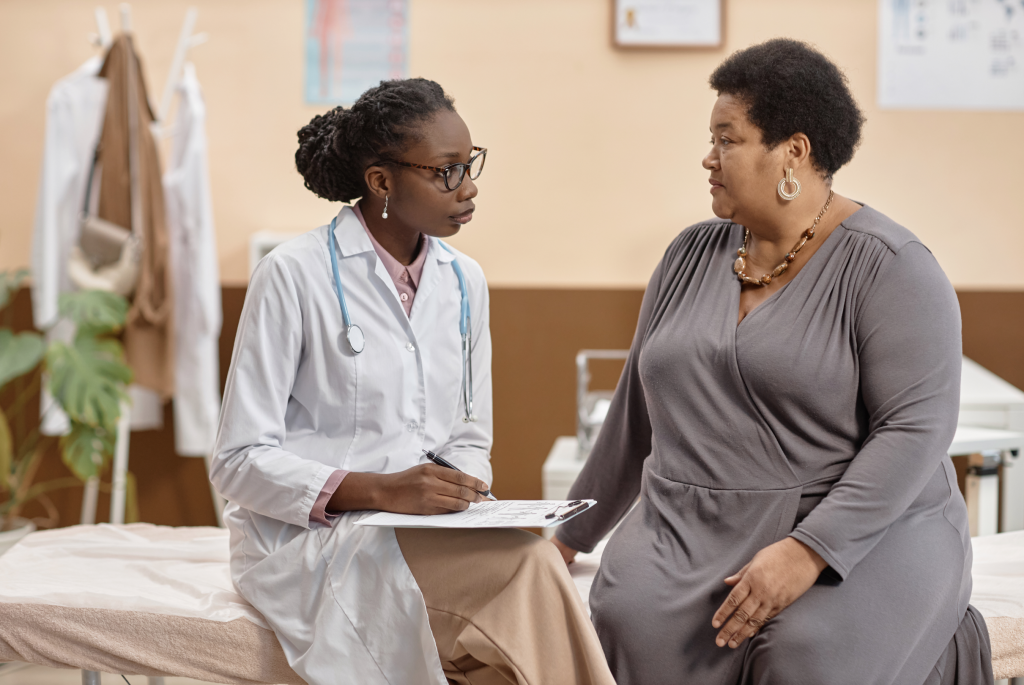 A doctor in a white
coat with a stethoscope sits
beside a patient on an exam
table, taking notes on a clip-
board. The patient, wearing
a gray dress, listens atten-
tively during a consultation
in a medical office.
